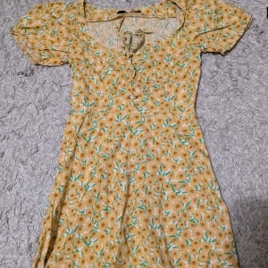 Gul blommig kortklänning BikBok XS - Supersöt gul kortklänning från BikBok i storlek XS med blommigt mönster, puffärmar och snörning vid urringningen. Perfekt för sommarens alla tillfällen och har en lätt och luftig känsla.
