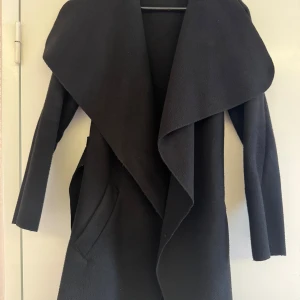 Svart omlott kappa med bälte - Snygg svart kappa med bred krage och omlott-design. Jackan har ett matchande bälte i midjan och två fickor framtill. Långärmad och med en stilren, loose passform som ger en chill vibe. 