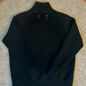 Svart zip tröja från Maison Margiela - Riktigt snygg svart ribbstickad kofta från Maison Margiela med hög krage och silverfärgad dragkedja. Ikoniska vita stygn i nacken och breda muddar. Perfekt för dig som gillar stilrena och exklusiva plagg med edge.