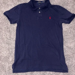 Marinblå pikétröja Polo Ralph Lauren - Snygg marinblå pikétröja från Polo Ralph Lauren med röd broderad logga på bröstet. Slim fit-modell med klassisk krage och två knappar framtill. Tillverkad i mjuk bomull, perfekt för en clean och stilren look.
