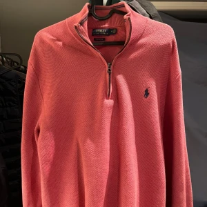 Rosa half zip tröja från Polo Ralph Lauren - Snygg rosa långärmad tröja från Polo Ralph Lauren med half zip och klassisk broderad logga på bröstet. Tröjan är stickad i bomull och har en avslappnad passform som är perfekt för chill dagar. Perfekt att slänga på sig när det är lite kyligt ute.