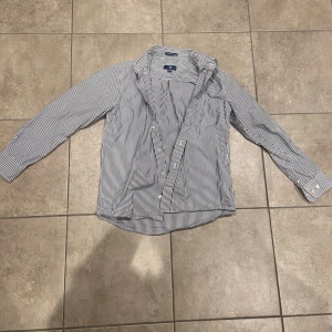 Randig skjorta från GANT, 146/152 - Snygg skjorta från GANT i klassiskt blåvitt randigt mönster. Skjortan har lång ärm, bröstficka och knappar framtill. Tillverkad i mjuk bomull som känns skön mot huden. Perfekt för dig som gillar stilren och tidlös look.