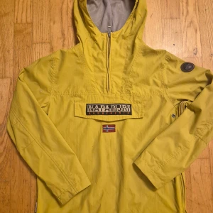 Napapijri - Snygg gul vindjacka från Napapijri med huva och halv dragkedja. Jackan har ett stort Napapijri-märke på bröstet, norsk flagga och en patch på ärmen. Tillverkad i polyester, perfekt för blåsiga dagar. Långärmad och loose passform.