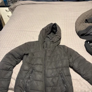 Svart pufferjacka från FB Sister - Svart pufferjacka från FB Sister med huva och två sidofickor med dragkedja. Jackan har quiltad design och hel dragkedja framtill. Storlek S.