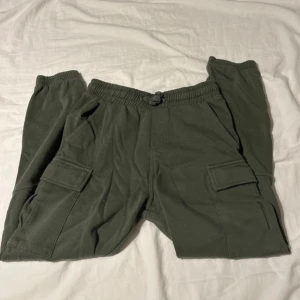 Gröna cargopants med snörning - Säljer ett par gröna cargopants med elastisk midja och snörning. Byxorna har flera fickor med lock på sidorna och baktill, samt muddar vid bensluten för en chill look. Materialet är mjukt och känns som bomull, perfekt för en avslappnad stil.
