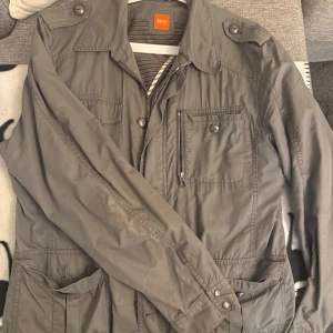 Khakifärgad jacka från Hugo Boss - Snygg khakifärgad tunnare jacka från Hugo Boss med flera fickor framtill, axelklaffar och dragkedja. Jackan har en klassisk krage och knappar på ärmsluten. Perfekt för dig som vill ha en stilren och funktionell jacka med coola detaljer.