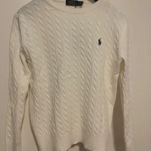 Vit  Polo Ralph Lauren - Vit kabelstickad tröja från Polo Ralph Lauren med klassisk rund halsringning och broderad logga på bröstet. Tröjan har lång ärm och ribbade muddar. Perfekt för lager-på-lager och snygg till jeans eller kjol.