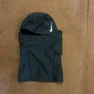 Nike Pro Therma-FIT Hood svart - Nike Pro Therma-FIT Hood i svart med vit Nike-logga. Tillverkad i ett syntetmaterial som är designat för att hålla värmen och transportera bort fukt. Huva med tight passform som täcker hela huvudet och halsen, perfekt för träning utomhus. Märkt med Nike Pro-etikett.