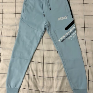 Ljusblå Hoodrich mjukisbyxor M - Snygga ljusblå mjukisbyxor från Hoodrich med tryck på benet och svarta detaljer. Byxorna har snörning i midjan, ribbade muddar vid benslut och är tillverkade i 80% bomull och 20% polyester. Perfekt för chill eller streetwear-look.