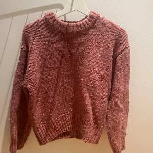 Rosa stickad tröja med grov struktur - Mysig stickad tröja i en dämpad rosa nyans med grov struktur och ribbade muddar vid hals, ärmslut och nederkant. Perfekt för lager-på-lager och chill dagar. Långärmad och loose fit för en avslappnad vibe.