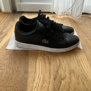 Svarta sneakers från Lacoste - Tja säljer ett par svarta Lacoste skor pga av ingen användning av de längre. Svarta sneakers från Lacoste med klassisk rund tå och vit platt sula. Skorna har snörning och är tillverkade i skinn med den ikoniska gröna krokodilen broderad på sidan. Perfekt för dig som gillar stilrena och sportiga vibes.pris kan eventuellt diskuteras 