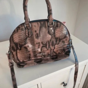 Brun handväska i ormmönstrat från Makgio - Snygg handväska från Makgio i brunt ormmönstrat material med både handtag och axelrem. Väskan har rundad form, dragkedja upptill och insida i klarblått tyg med flera fack. Perfekt för dig som vill sticka ut med en unik accessoar.