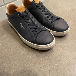  Pepe Jeans skor - Snygga mörkblå sneakers från Pepe Jeans med vita detaljer och logga på sidan och hälen. Skorna har klassisk snörning, rund tå och platt vit sula. Ovandelen är i skinn och insidan har en ljusbrun läderlook. Perfekta för en clean och stilren look. Har låda kvar, helt nya, har bara testat dem och de är för små för mig så säljer dem