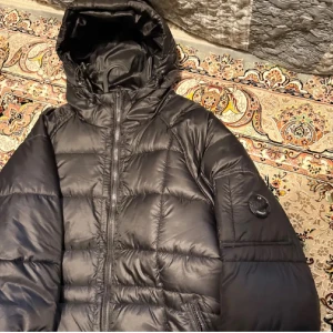 Svart dunjacka från C.P. Company - Säljer en svart dunjacka från C.P. Company med ikonisk linsdetalj på ärmen. Jackan har huva, dragkedja framtill och är tillverkad i glansig nylon. Perfekt för kalla dagar och har en modern, puffig look. Helt ny oanvänd för stor för mig, storlek L