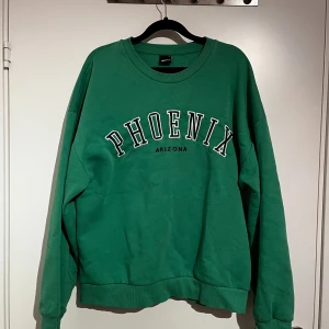 Grön sweatshirt med PHOENIX-tryck, Gina - Säljer en grön sweatshirt från Gina Tricot med stort vitt PHOENIX-tryck och svart ARIZONA-text på bröstet. Tröjan har rund hals, ribbade muddar och en loose passform.