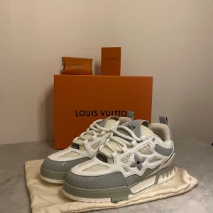 Louis Vuitton Skate sneakers vit/grå - Louis Vuitton Skate sneakers i vitt och grått med chunky platt sula, snörning och LV-blomdetalj på sidan. Mesh och skinn i ovandelen, broderad 1984-logga på hälen och coola detaljer. Perfekt för dig som gillar exklusiv streetwear och snygg design.