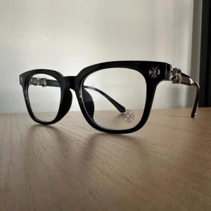 Svarta glasögon Chrome Hearts - Snygga svarta glasögon från Chrome Hearts med fyrkantig form och coola silverdetaljer vid gångjärnen. Bågarna har en stilren design med blommönster på sidorna och en unik blomdetalj på glaset. Perfekt för dig som vill sticka ut med en exklusiv look.