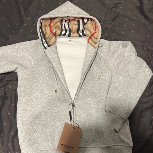 Grå hoodie från Burberry med dragkedja - Snygg grå hoodie från Burberry i junior storlek L men passar som S, med klassiskt rutigt foder i huvan och broderad logga på bröstet. Tröjan har hel dragkedja, snörning i huvan och känguruficka framtill. Perfekt för dig som vill ha en stilren och lyxig streetwear-look.