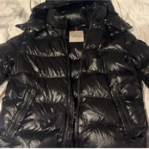 Svart pufferjacka från Moncler - Säljer en svart, glansig pufferjacka från Moncler med hög krage och två snedställda dragkedjefickor. Jackan har klassisk quiltad design och tydlig Moncler-logga på insidan. Perfekt för kalla dagar och riktigt snygg streetstil.