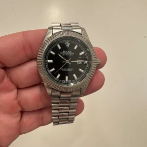 Silverfärgad klocka, rolle  - Silvrig klocka med svart urtavla och silverfärgat armband i metall. Klockan har en räfflad bezel, datumfönster med förstoringsglas och klassisk länkdesign. Snygg och tidlös modell med tydliga indexmarkeringar och logga på spännet. (Följer med extra länkar)