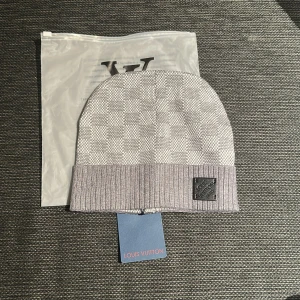 Grå rutig mössa  - Snygg grå mössa  med klassiskt rutigt mönster och ribbad kant. Mössan har en svart patch med  framtill och är tillverkad i ett mjukt och lyxigt material. Perfekt accessoar för att lyfta din vinterstil.