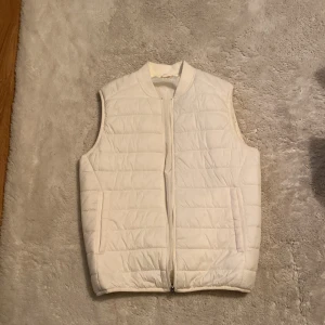 Vit quiltad dunväst från Zara - Säljer en stilren vit dunväst från Zara i storlek S. Västen är quiltad med horisontella sömmar, har dragkedja framtill och två fickor på sidorna. Perfekt att slänga över en hoodie eller tröja för en clean look.