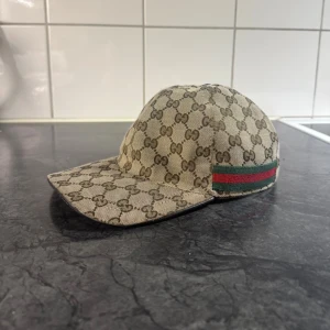 Gucci keps med GG-mönster - Säljer en beige keps från Gucci med klassiskt GG-monogram över hela kepsen. Kepsen har grönt och rött band på sidan och justerbar rem baktill i mörkbrunt skinn. Materialet är bomullscanvas och insidan är svart. Perfekt för dig som gillar lyxiga accessoarer.