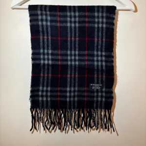 Rutig halsduk i kashmir från Burberry - Snygg rutig halsduk från Burberry i 100% kashmir. Mörkblå bas med grå och röda rutor samt fransar i ändarna. Perfekt accessoar för att lyfta din outfit med klassisk brittisk stil.