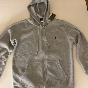 Grå hoodie från Polo Ralph Lauren - Snygg grå hoodie från Polo Ralph Lauren med dragkedja framtill och klassisk huva med snörning. Tröjan har två stora fickor fram och den ikoniska lilla loggan broderad på bröstet. Perfekt för chill dagar eller när du vill ha en enkel och stilren look.
