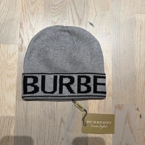 Grå Burberry mössa med logga - Snygg grå mössa från Burberry med bred svart kant där BURBERRY är stickat i stora bokstäver. Klassisk form och mjukt stickat material som sitter skönt på huvudet. Perfekt för dig som vill ha en stilren accessoar med lyxig känsla.
