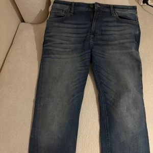 Blå jeans från Jack & Jones, storlek 33/32 - Snygga blå jeans från Jack & Jones med klassisk femficksdesign och diskreta slitningar framtill. Jeansen har rak passform och är tillverkade i robust denim med en mörkblå tvätt. Perfekt för dig som gillar en stilren och avslappnad look.