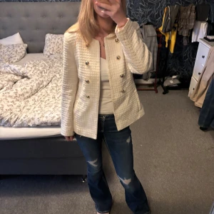 Beige kavajjacka från Zara med guldknappar - Stilren beige kavajjacka från Zara med strukturerat tyg och dekorativa guldknappar framtill. Jackan har rund halsringning, raka långa ärmar och två fickor. Perfekt för dig som vill ha en klassisk men trendig look.