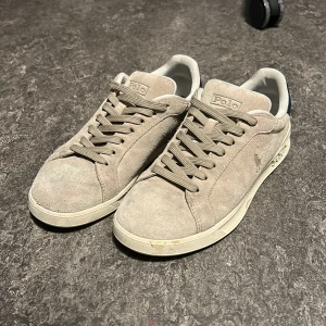 Beige mockasneakers från polo ralph - Snygga sneakers i beige mocka från Polo Ralph Lauren med klassisk låg profil. Skorna har snörning, vit sula och mörkblå detalj med Polo-logga på hälen. Perfekta för en clean och stilren look.