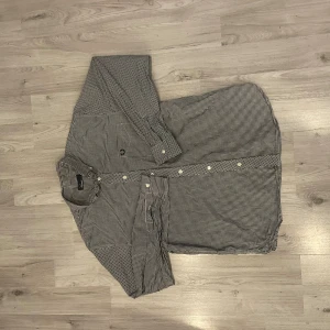 Svartvit rutig Fred Perry skjorta L - Snygg svartvit rutig skjorta från Fred Perry i storlek L. Skjortan har klassisk krage, vita knappar och det ikoniska Fred Perry-logot på bröstet. Tillverkad i 100% bomull för skön känsla och bra passform. Perfekt för dig som gillar stilrena och tidlösa plagg.