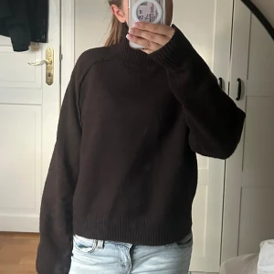Mörkbrun stickad polotröja - Säljer en mörkbrun stickad polotröja med hög krage och långa ärmar. Tröjan har en loose passform och ribbade muddar vid ärmslut och nederkant. Perfekt för kyliga dagar och enkel att styla med jeans eller kjol.