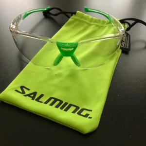 Salming sportglasögon med gröna detaljer - Säljer ett par sportiga glasögon från Salming med transparent båge och coola gröna detaljer vid näsan och skalmarna. Perfekt för dig som vill ha skydd och stil på träningen. Kommer med en neongul Salming-påse för förvaring.