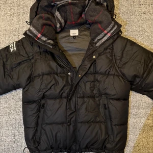 Svart pufferjacka från Burberry L - Svart pufferjacka från Burberry med huva och klassiskt rutigt foder i grått, vitt och rött. Jackan har dragkedja, knappar och två fickor med dragkedja framtill. Perfekt för kalla dagar och man kan även göra om den till en vest om man vill det