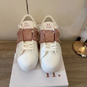 Valentino Rockstud sneakers vit/rosa - Säljer ett par Valentino Rockstud sneakers i vitt skinn med ljusrosa detaljer och ikoniska nitar. Skorna har snörning, platt sula och rund tå. Snygg kontrast mellan det vita och rosa, och nitarna ger en edgy vibe. Perfekta för dig som gillar designer och vill sticka ut.
