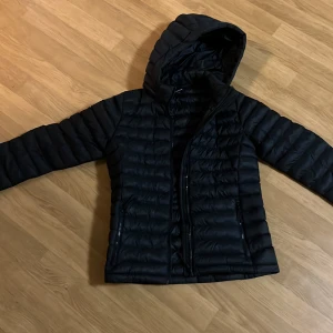 Svart dunjacka från Everest med huva - Svart dunjacka från Everest med snygg quiltad design och praktisk huva. Jackan har dragkedja framtill och två sidofickor med dragkedja. Perfekt för kalla dagar och har en stilren look som funkar till det mesta.