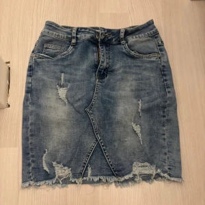 Blå jeanskjol med slitningar från Newplay - Snygg blå jeanskjol från Newplay med slitna detaljer och fransad nederkant. Kjolen har klassisk femficksdesign, bälteshällor och omlott-effekt framtill. Perfekt för dig som gillar en cool och avslappnad stil. Pris kan diskuteras!