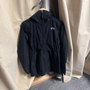 Svart vindjacka från The North Face - Svart vindjacka från The North Face med huva och hel dragkedja framtill. Jackan har två stora fickor, justerbar midja och logga på bröstet och ryggen. Perfekt för blåsiga dagar och enkel att matcha med allt.