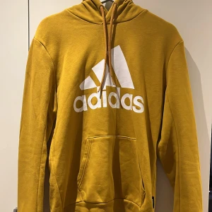 Gul Adidas hoodie med logga - Snygg gul hoodie från Adidas med stor vit logga på bröstet. Tröjan har huva med snörning, magficka och ribbade muddar. Perfekt för chill eller sportiga dagar. Tillverkad i mjuk bomullsmix som känns skön mot huden.