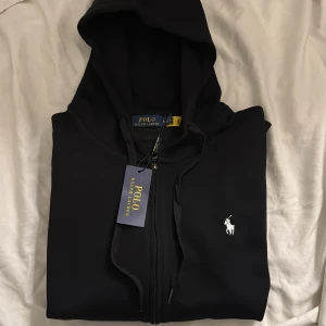 Svart hoodie från Polo Ralph Lauren - Svart hoodie från Polo Ralph Lauren med klassisk vit logga broderad på bröstet. Tröjan har huva med snörning och dragkedja framtill. Mjuk bomullskvalitet och stilren design som funkar till jeans eller joggers.