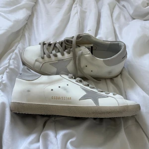 Golden Goose Superstar  - Golden Goose Superstar sneakers i vitt skinn med grå detaljer och snörning. Ikonisk stjärna på sidan och GGDB/SSTAR-tryck i guld. Platt sula med svart undersida och rund tå. Grå hälparti med Golden Goose-logga. Perfekt för dig som gillar clean och stilren look. Og ingår och kvitto finns! Tveka inte på att höra av dig🙌