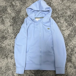 Ljusblå hoodie från Lacoste - Snygg ljusblå hoodie från Lacoste i klassisk passform. Tröjan har huva med snörning, dragkedja framtill och två fickor. Ikoniska Lacoste-krokodilen är broderad på bröstet. Perfekt för chill dagar och enkel att matcha med jeans eller joggers.