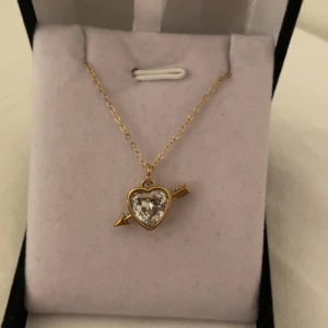 Halsband med hjärta och pil i guld - Superfint halsband med tunn guldfärgad kedja och ett hängsmycke i form av ett hjärta med en pil genom. Hjärtat är dekorerat med en glittrande, klar sten som ger extra bling. Perfekt accessoar för att lyfta vilken outfit som helst. Rostfri