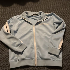 Ljusblå track jacket från Palm Angels - Snygg ljusblå track jacket från Palm Angels med vita och svarta ränder längs ärmarna. Jackan har dragkedja framtill, ribbade muddar och klassisk sportig look. Perfekt för dig som gillar streetwear och vill sticka ut med en fräsch färg.