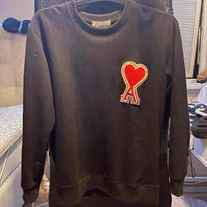 Ami paris sweatshirt - Svart sweatshirt från Ami med stor röd hjärtpatch på bröstet. Tröjan har rund halsringning, ribbade muddar och är tillverkad i mjuk bomull. Perfekt för dig som gillar stilrena plagg med en twist.