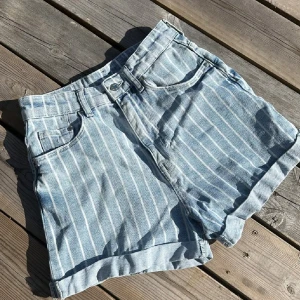 Randiga ljusblå jeansshorts - Säljer ett par ljusblå jeansshorts med vita vertikala ränder. De har hög midja, klassiska fickor fram och bak samt uppvikta benslut. 