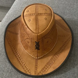 Brun cowboyhatt i skinn med mönster - Säljer en brun cowboyhatt i skinn med snygga ingraverade mönster och kontrasterande svart kant. Hatten har dekorativa sömmar och ett coolt snörningsdetalj framtill. Perfekt för dig som vill sticka ut med westernstil.
